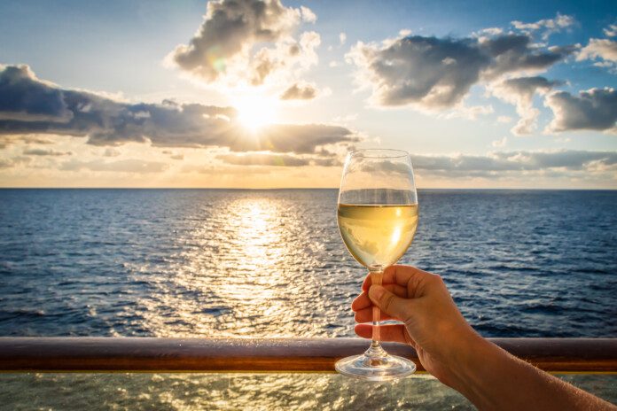 During sunset a female hand toasts the new year with a glass of white wine. Photo taken on a balcony of a cruise ship with a sea view. Cloudy sky.