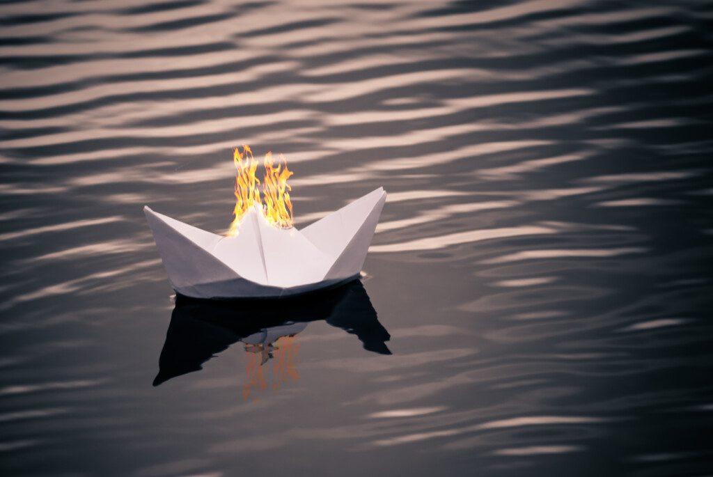 Small paper ship floating on water. Fire on the boat. Origami paper boat sailing on river with ripple (©iStock.com/art-sonik)