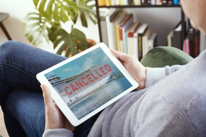 closeup of a young caucasian man, lying comfortably on an armchair, watching his tablet, with a picture of a cruise ship and the text cancelled in its screen, depicting the cancellation of a trip